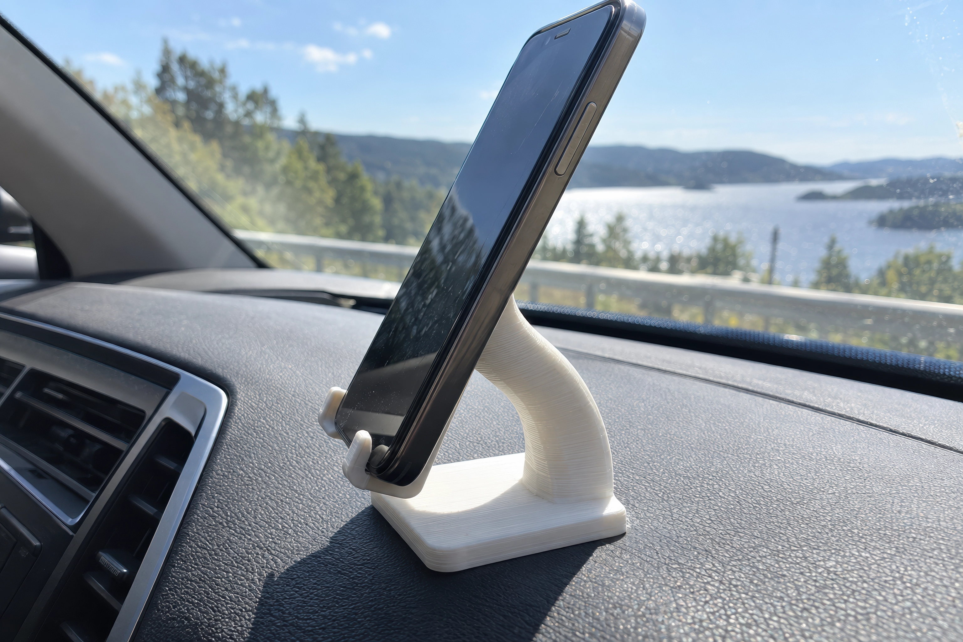 Et 3D-printet telefonstativ i hvit PLA på dashbordet i en bil i sterk sol, der armen har bøyd seg fremover av varmen.